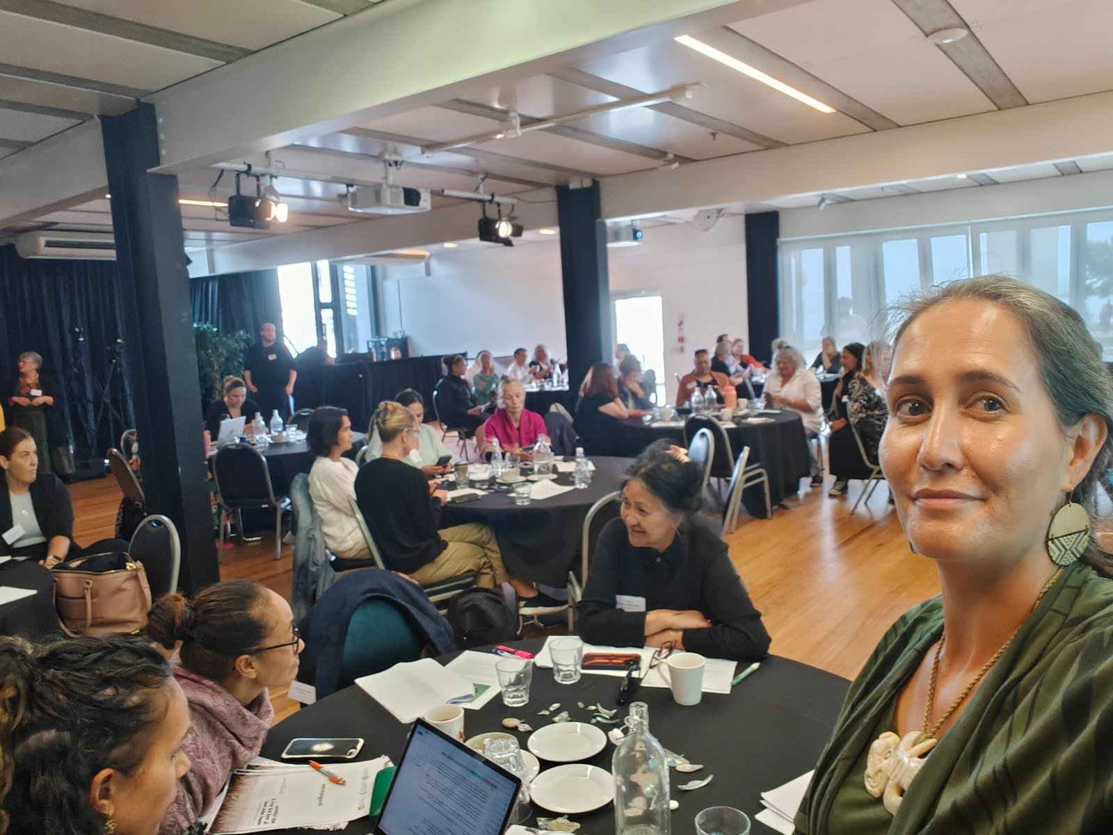 Maori woman taking a selfie of her and the the conference room behind her with people sitting at round tables
